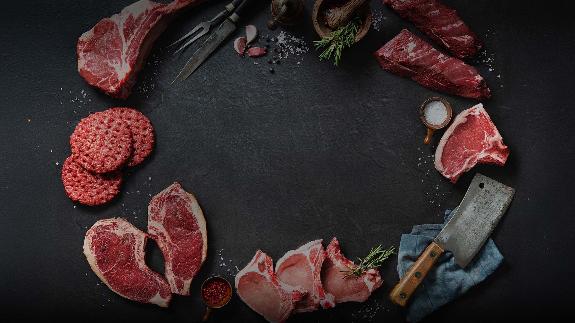 Karnivore Krate delivers Various raw cuts of red meat, burgers, pork chops, herbs, spices, salt, a meat cleaver, and a knife are arranged in a circle on a dark surface, leaving an empty space in the center.