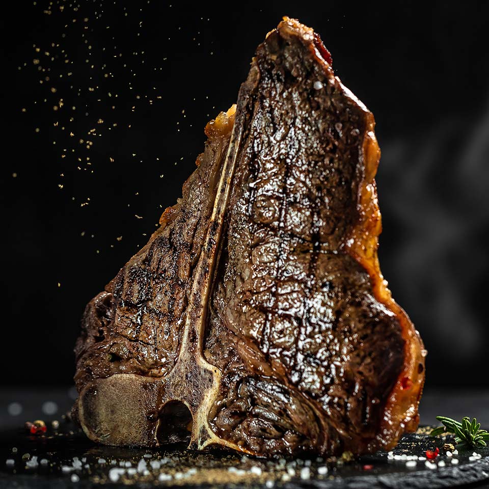 Grilled juicy t bone steak sits up right and is sprinkled with coarse salt and pepper.