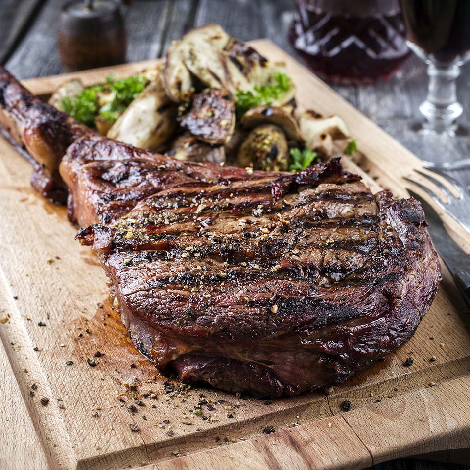 Juicy grilled t bone beef steak rests on wood cutting board served with a side of grilled potatoes and mushrooms.