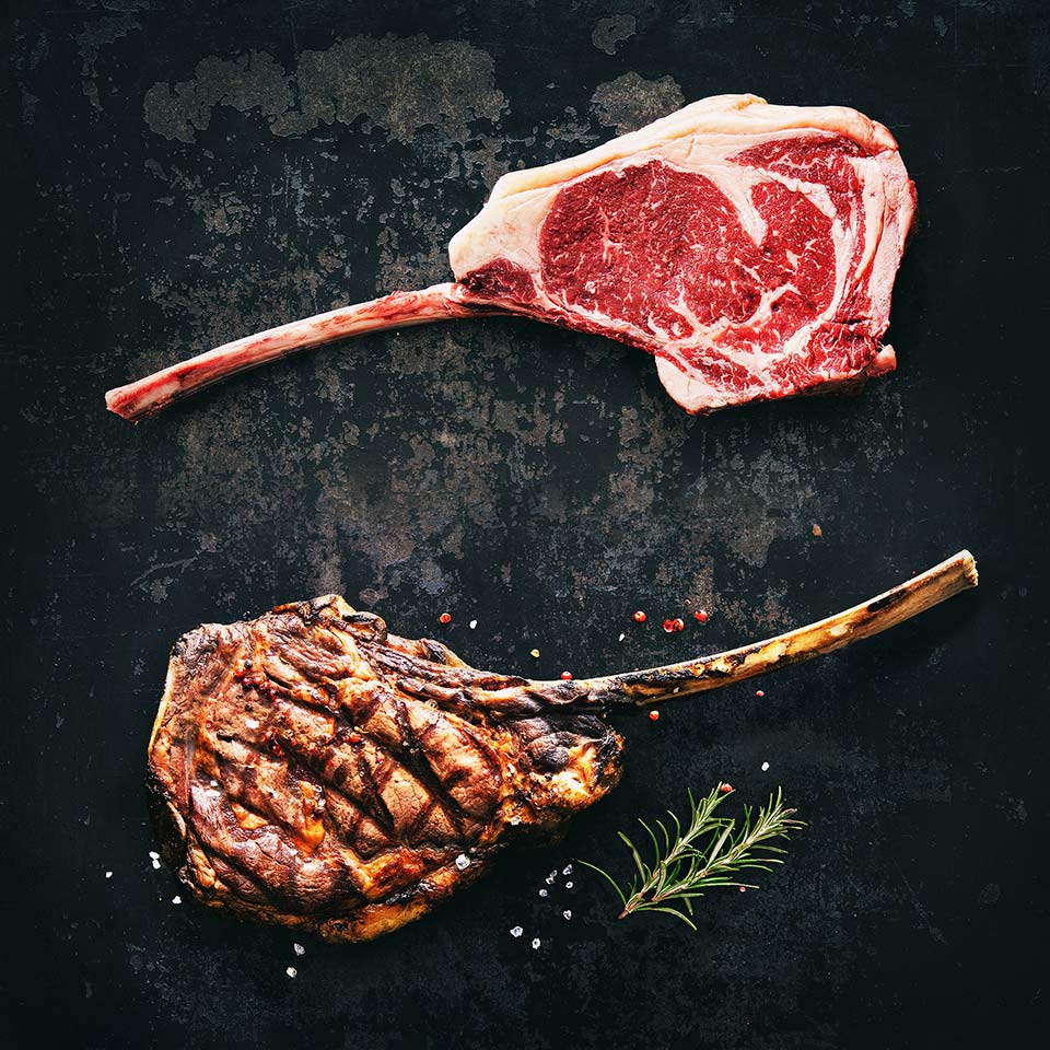 Two Tomahawk steaks, top steak is raw, bottom steak is grilled and seasoned with pepper, salt and rosemary.