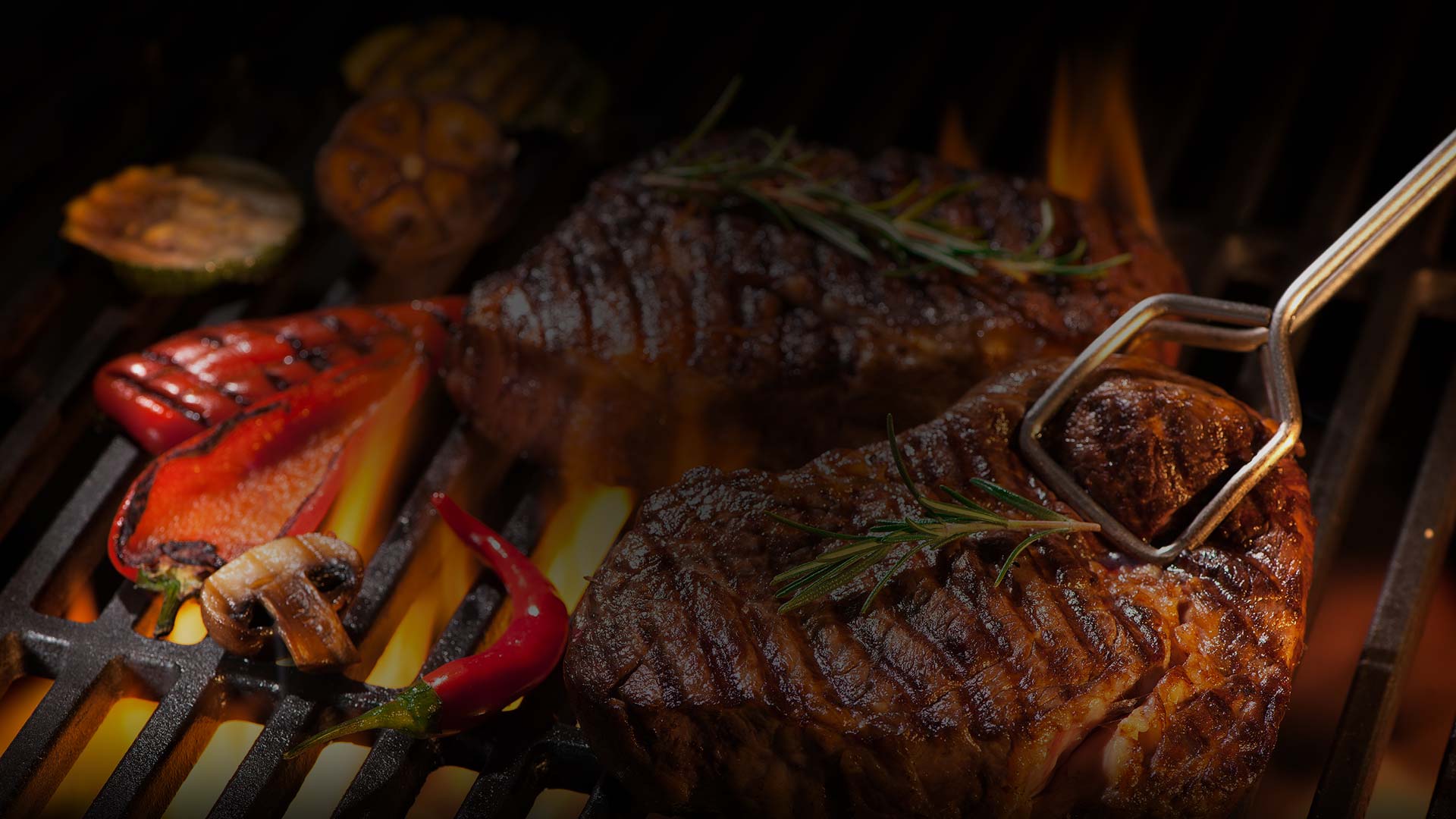 Two grilled steaks with rosemary sizzle on a barbecue grill, surrounded by veggies. Enjoy this scene at home with Steaks Delivered from Karnivore Krate, your meat subscription box for premium cuts and delicious meals.