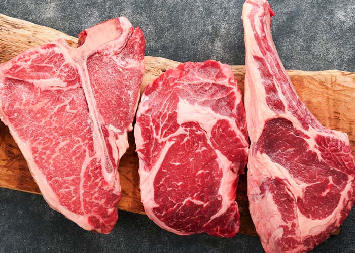 Three raw cuts of red meat, including a T-bone, ribeye, and other bone-in steaks, are arranged on a wooden board against a dark gray background—perfect selections from Karnivore Krate Premium Meat Subscription Boxes delivered nationwide.