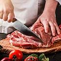 A person in a white chef’s coat slices premium Angus meat on a wooden cutting board, surrounded by fresh vegetables such as tomatoes and bell peppers.