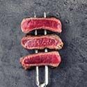 Three slices of premium meat, seared outside and pink inside, are stacked on a two-pronged fork against a dark, textured backdrop—perfect for showcasing your next meat subscription box discovery.